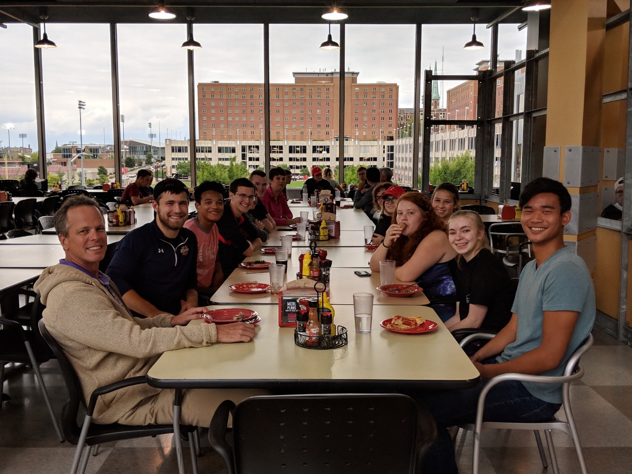 about 15 students and faculty members sit around a table eating pizza.