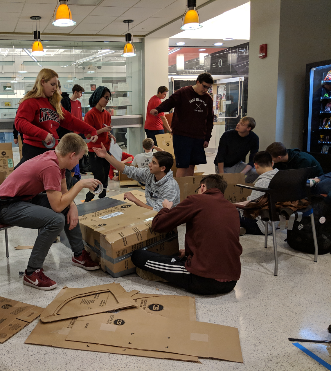 three groups of about a dozen in casual dress, stand and sit in a common area filled with cardboard and chairs.