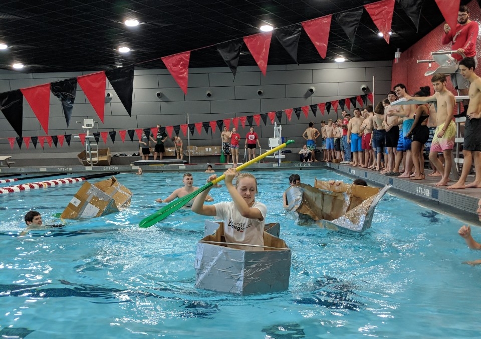 Several students in bathing suits compete in a cardboard canoe race in a UC pool. In the foreground, one young woman is paddling her canoe towards the camera, while two other canoes sink in the background. Groups of people stand on the side of the pool watching.